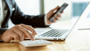 Un homme en costume utilise une calculatrice tout en consultant un smartphone, devant un ordinateur portable. litiges