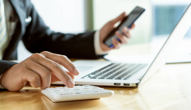 Un homme en costume utilise une calculatrice tout en consultant un smartphone, devant un ordinateur portable.