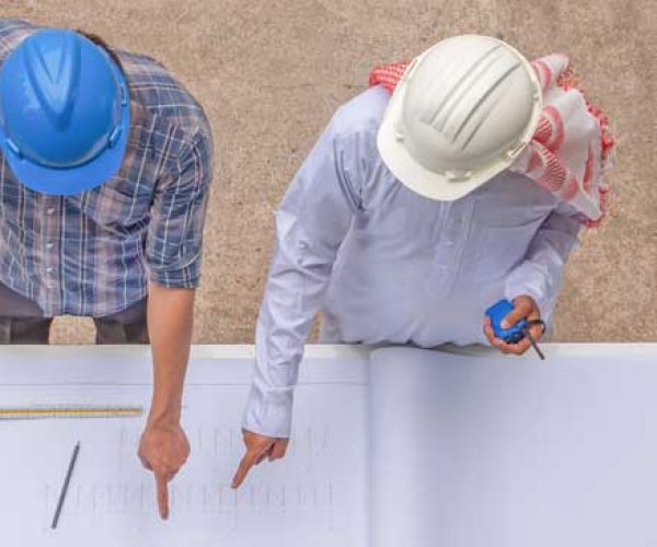 AMOA Deux assistants à maîtrise d'ouvrages, portant des casques de sécurité, analysent des plans de construction sur une table.