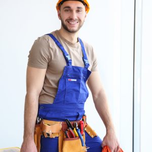 Young smiling electrician holding bunch of wires and tool box indoors Un artisan au travail, illustrant la raréfaction des artisans français et les défis du métier.